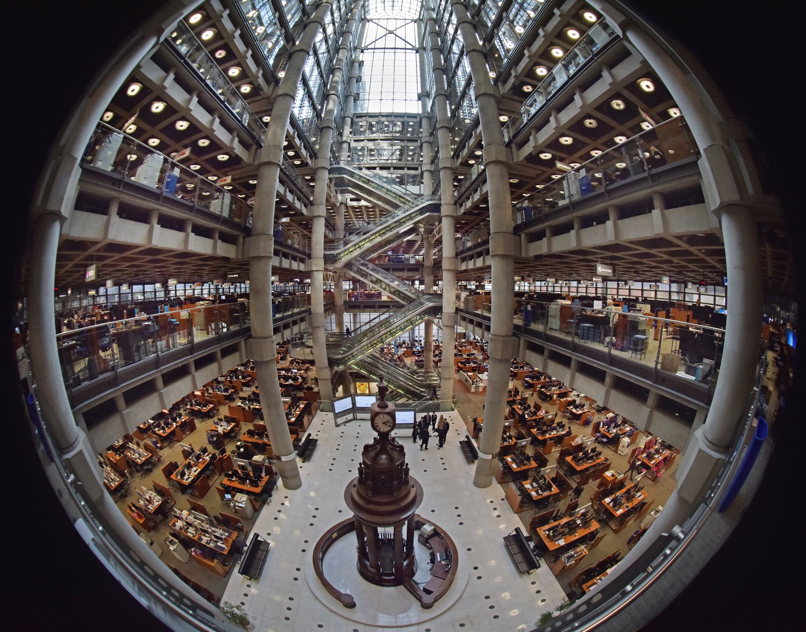 Fisheye photograph looking down into a vast atrium with a glass roof, escalators and exposed concrete columns, and rows of wooden desks on the floor around a central clock and bell stand.